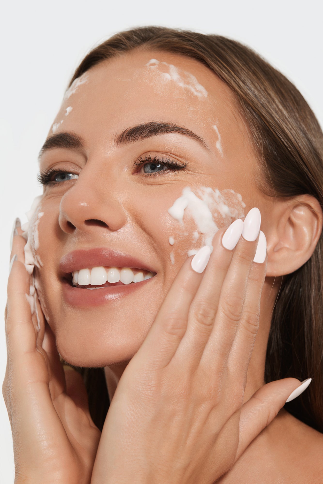 A smiling person applies foamy cleanser to their face with both hands.